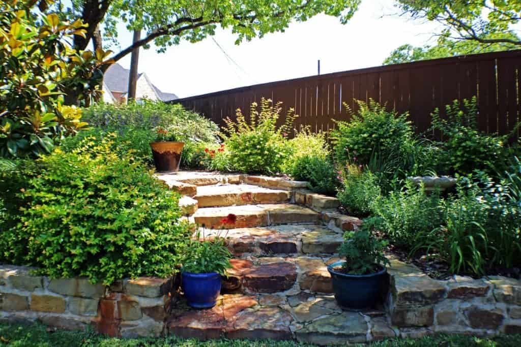 Stone steps lead up through a lush garden with green shrubs, potted plants, and a wooden fence, an inviting example of hardscaping Tarrant County homeowners love.