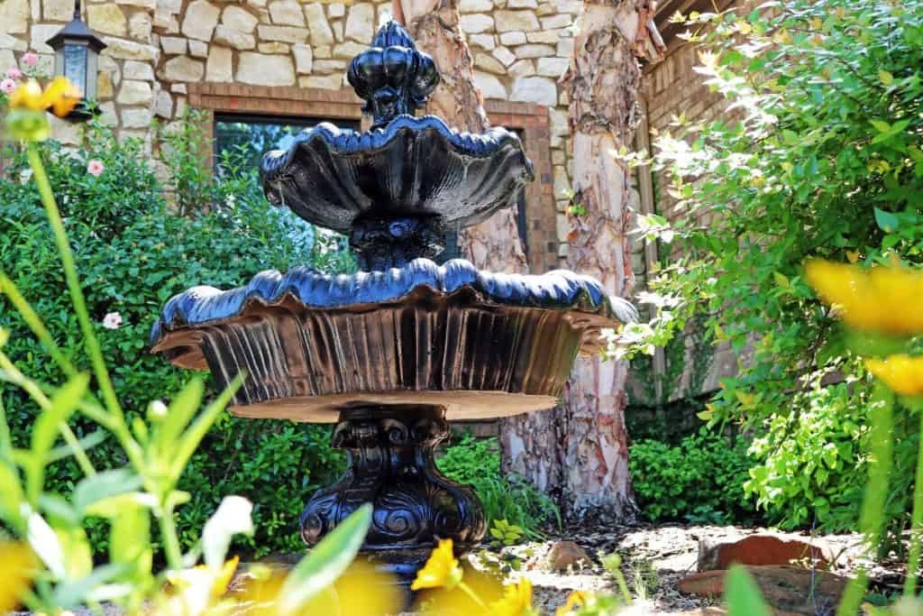 Black two-tiered fountain in a garden surrounded by green bushes, yellow flowers, and stone walls, an elegant example of landscape water features enhancing outdoor spaces in Tarrant County.