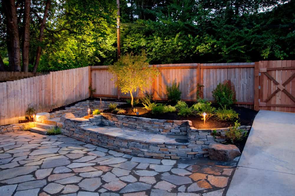 A backyard in Tarrant County featuring hardscaping with a stone patio, raised garden bed, small shrubs, wooden fence, and inviting outdoor lighting at dusk.